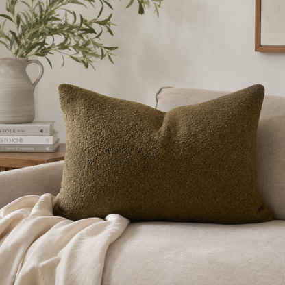 An olive green bouclé cushion resting on a light beige sofa with a cream throw blanket and a ceramic pitcher with greenery in the background.