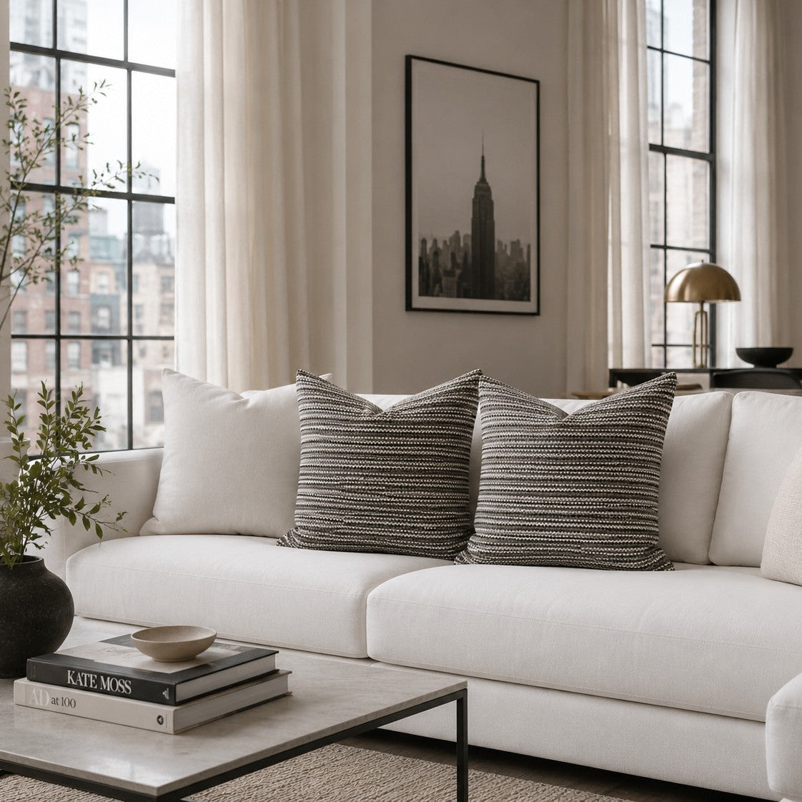 A modern living room scene featuring a white sectional sofa with two textured gray and white striped scatter cushions. The room has large windows with sheer curtains, a black-framed cityscape print on the wall, and a glass coffee table in the foreground.