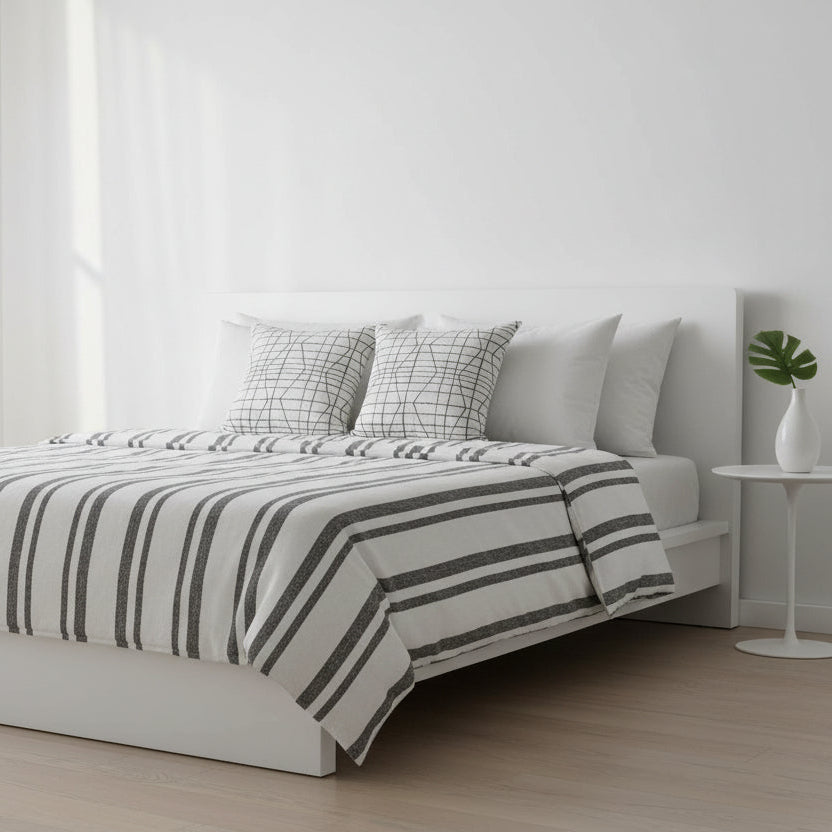 Bedroom with a bed featuring a striped duvet and patterned scatter cushions, white curtains, and a small table with a plant.
