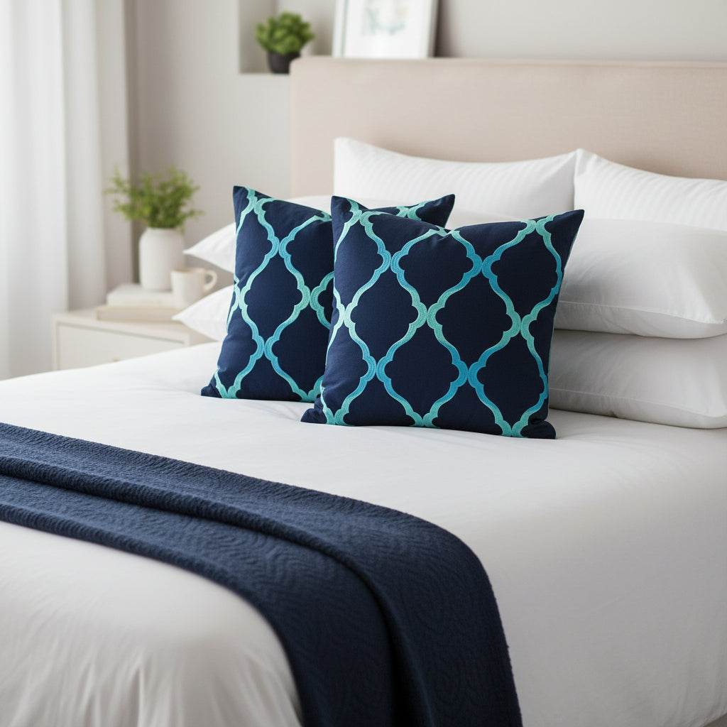 Two navy blue cushions with a turquoise geometric pattern on a bed dressed in white bedding with a navy throw blanket 
