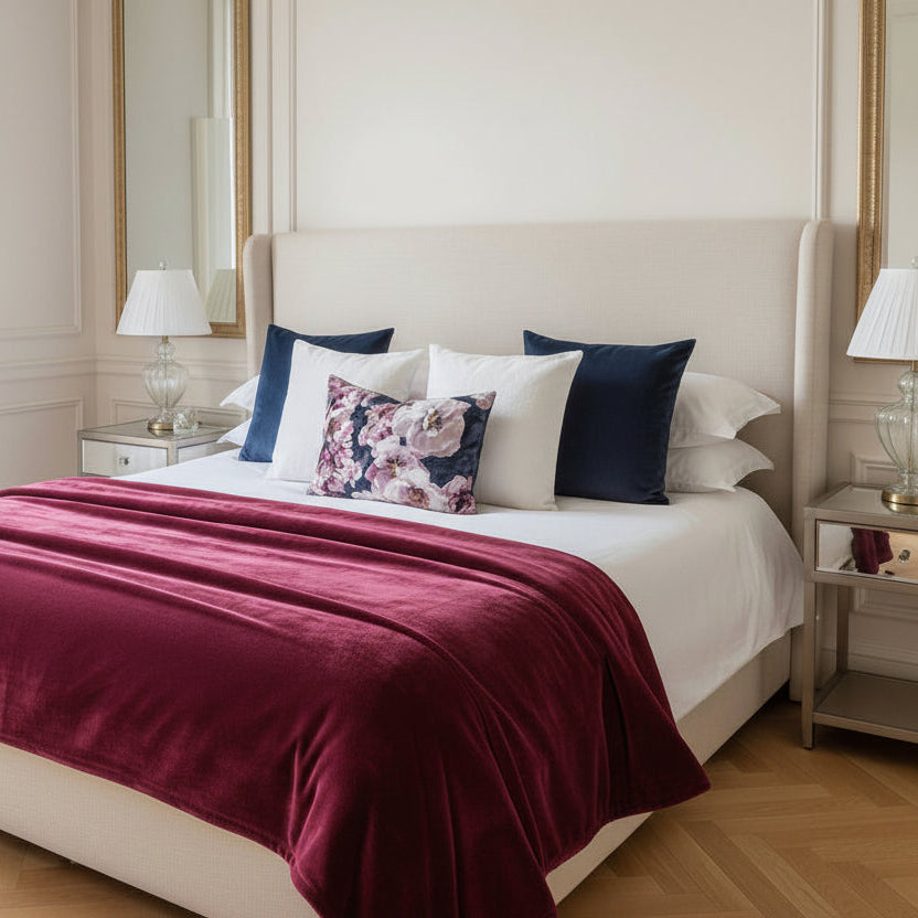 Neatly made bed with decorative cushions and a deep pink throw blanket in a well-lit bedroom.